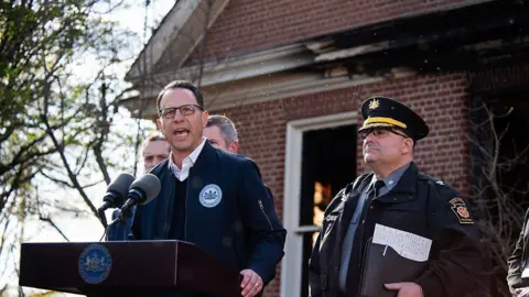 Getty Images Governor Josh Shapiro speaks at a podium in front of a brick home with fire damage; he's wearing a jacket and glasses.