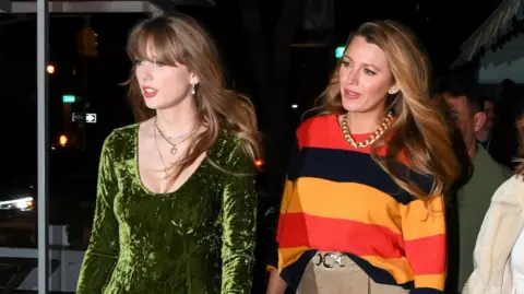 Getty Images Taylor Swift, wearing a green velvet dress, walks with Blake Lively, wearing a black, red and orange jumper, at night in New York City in 2023.