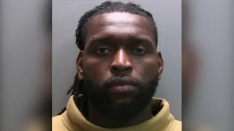 NCA Custody photo of Emmanuel-Thomas. He looks serious and is wearing a yellow turtle neck jumper.