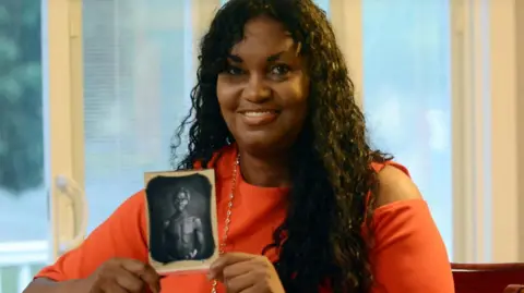 Reuters Tamara Lanier wearing a red dress holds a copy of an black and white 1850 photo of a man she says is her enslaved ancestor "Papa Renty" Taylor in Norwich