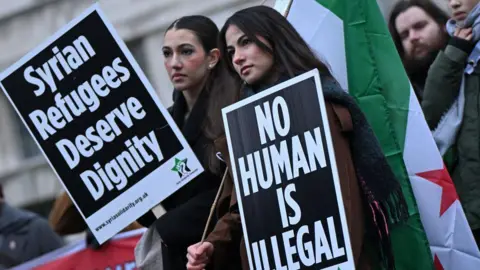AFP via Getty Images Two women stand outside 10 Downing Street holding  Syrian flag and placards reading: 'Syrian refugees deserve dignity' and 'no human is illegal'. 