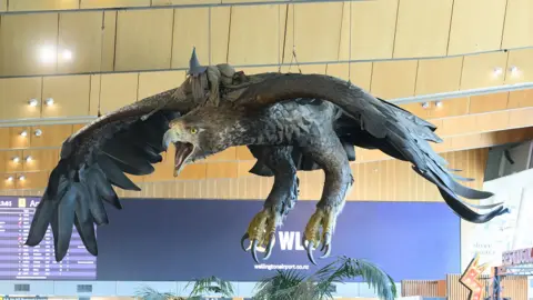 Wellington Airport Side view of the giant eagle hanging from the ceiling, with Gandalf riding on its back. Behind the sculpture is a large purple screen showing flight arrival and departure times.