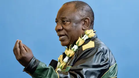 AFP/Getty Images Cyril Ramaphosa, wearing a leather green, yellow and black leather jacket and with a yellow and white rose garland around his neck, gestures with his hand as he talks.