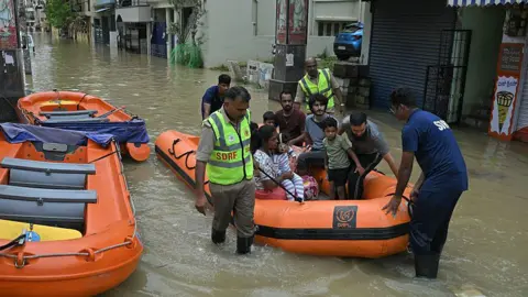 Getty Images State Disaster Response Force personnel rescue residents from a flooded locality following heavy rainfall in Bengaluru on May 19, 2025. The image shows people in orange rescue boats, floating through a flooded road.