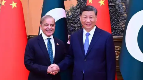 X/Shehbaz Sharif Pakistani President Shehbaz Sharif shakes hands with Chinese President Xi Jinping as the leaders smile while standing in front of the national flags of China and Pakistan.
