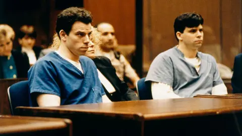 Getty Images The Menendez brothers sit in prison suits inside a courtroom. In between them, their defence attorney sits at the wooden table.