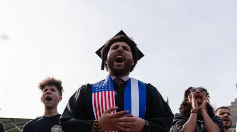 Bloomberg via Getty Images A Harvard University student chants with fellow demonstrators during a protest in support of international students at Harvard University.  He is wearing a graduation cap and gown with a sash which has an American flag on it. 