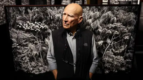 Getty Images Sebastião Salgado standing in front of a black and white photo of trees in the Amazon