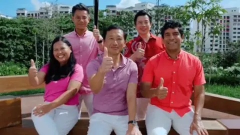 Ong Ye Kung/TikTok A screenshot of a TikTok video by Ong Ye Kung, showing him and four other PAP members wearing white pants and red or pink shirts. They are sitting on a park bench and giving the thumbs-up sign.