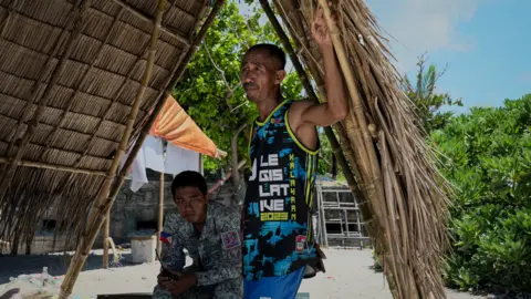 BBC/VIRMA SIMONETTE Two men stand in the shade of a shelter made of reeds