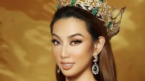 Miss Grand International Nguyen Thuc Thuy Tien smiles at the camera wearing an elaborate gold and green crown and jewelled earrings