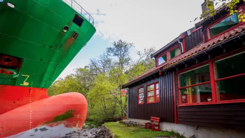 NTB/Jan Langhaugvia via Reuters The bow of a green and red container ship next to a small wooden cabin