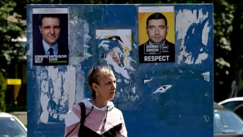 EPA-EFE/Shutterstock A woman walks past a billboard displaying posters of presidential candidates Nicusor Dan (left) and George Simion (right) in Bucharest, Romania. Photo: 15 May 2025