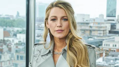 BBC Blake Lively is wearing a grey jacket and stood with a London skyline behind her.