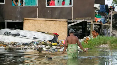 Getty Images A woman wrapped in a green patterned sarong wades through knee-deep water to reach her home. The house is on stilts and surrounded by rubbish. Washing hangs off the side of the building, and a small tin dinghy is upside-down beside the building. 