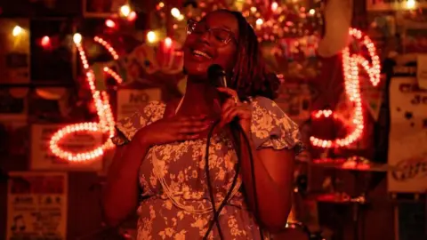 Reuters/ Kevin Wurm Edna Nicole Luckett sings at a microphone, her hand on her heart, with light-up signs showing musical notes behind her
