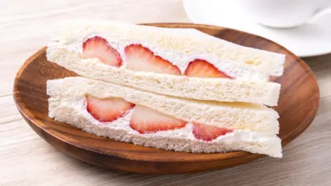 Promo_Link/Getty Images A strawberry and cream sandwich in white bread, served on a wooden plate on top of a table