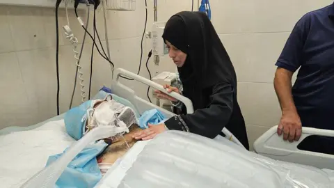 Getty Images Hamdi al-Najjar is visited by his niece, Samah Al-Najjar, while in the intensive care unit. His face is heavily bandaged and he is lying in a hospital bed. His niece is dressed all in black, has he hand on his chest and is visibly upset.