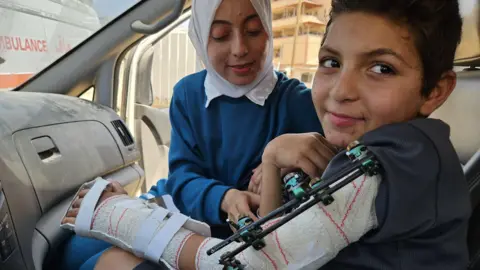 Getty Images Adam al-Najjar sits in an ambulance with a brace around his arm wearing a dark t-shirt while his cousin, wearing blue and a white headscarf, tends to him.