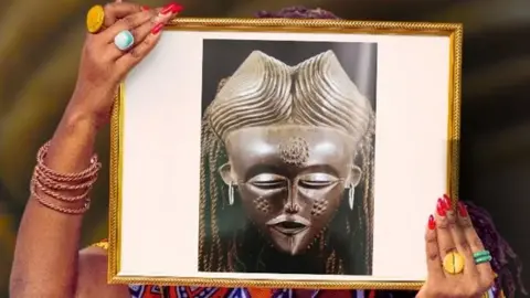 Women’s History Museum Zambia Samba Yonga from Women's History Museum of Zambia holds up a frame over her face showing a photograph of a sacred mask with Sona symbols etched on to its surface, each telling stories of women's significance, wisdom, and the vital knowledge they carried.