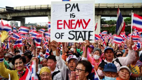 Reuters A woman standing in the centre of a crowd that fills the image, holding a placard that reads "PM is enemy of state" in black lettering, with enemy written in red. the entire crowd is filled of people waving the Thai flag. In the background is an overpass.