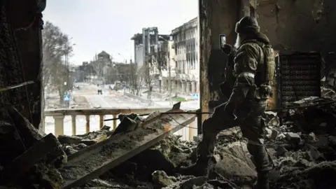 Getty Images A Russian soldier stands in the ruins of Mariupol's theatre in spring 2022, taking a photo out of a destroyed window using his phone