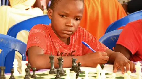 Robert Katende A child in a red T-shirt holding a pen concentrates as they look at a chess board with pieces in play. Other young players are
