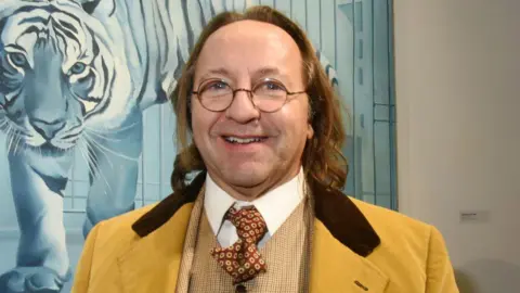 Getty Images Bill Pallot smiles in front of a painting at a gallery wearing round-rimmed glasses and a three-piece yellow suit.