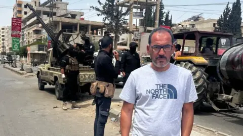 BBC Feras Kilani stands on a street in Damascus, Syria. In the background, Syrian security forces are deployed with a truck-mounted anti-aircraft gun (7 May 2025) 