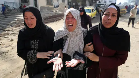Reuters Three women linking arms with expressions of despair, with woman in the centre crying. Woman on left of image is wearing a black abaya with a black hijab, woman in centre, the same with a white printed hijab and the woman on the right is wearing a burgundy abaya with a black hijab and white bandana underneath it.