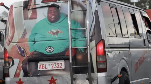 BBC A minibus taxi in traffic in Harare with its back painted with a big portrait of Sir Wicknell in a green long-sleeve top with polo players on its front. The vehicle's side panel has the words 'Sir Wicky' painted on it in red.