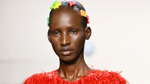 Getty Images A close-up of Arop Akol in a red plastic feathered outfit at the Ashish show during London Fashion Week in February 2025. Her hairs is decorated with plastic alphabet letters.