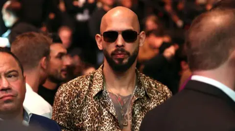 Getty Images Andrew Tate wearing dark sunglasses and a button-down animal print shirt and a dogtag necklace