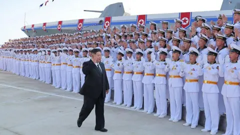 KCNA via Reuters Rows of soldiers in white uniforms and white caps cheer as Kim Jong Un walk in front of them, wearing a black suit and raising his hand. Behind them is a massive grey ship.