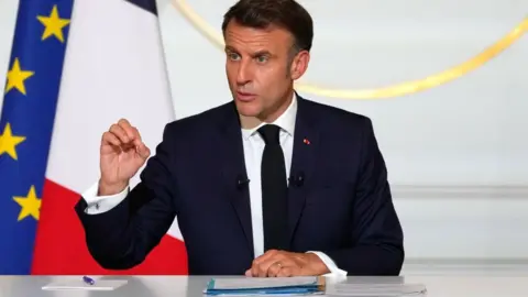 Michel Euler/Pool via Reuters Emmanuel Macron, wearing a dark suit, sits behind a desk at a conference, in front of an EU and a French flag