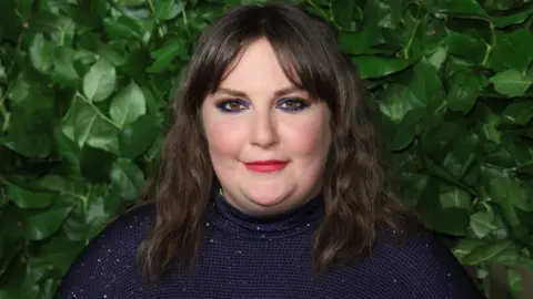Getty Images Lena Dunham - a woman with black hair and red lipstick - smiles at the camera wit a backdrop of foliage