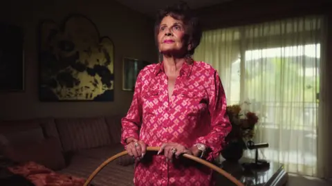 Chris Riess and Amy Hill Photo shows Joan Anderson in a bright pink button down shirt holding a wooden hoola hoop. She is glancing up at the sky and wearing pink lipstick with brown coiffed hair. She is stood in a room with curtains drawn and a striped sofa in the background.