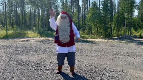 BBC/Erika Benke A man dressed as Santa waves at the camera. He is standing on concrete and surrounded by pine trees but the landscape in snowless