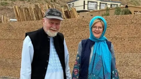 PA Media Peter and Barbie Reynolds smile at the camera in front of an orange brick wall. Peter is an elderly man wearing a blue long-sleeved top, a black waistcoat and a grey cap, while Barbie wears a bright blue headscarf, a navy gilet and a patterned blue top.