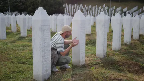 BBC A recent photograph of a man in a modern outfit (striped t-shirt, cargo trousers and a backwards cap) prays with his palms facing upwards next to a plain white grave. The cemetery is filled with thousands of identical graves where victims of the Srebrenica massacre are buried.