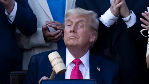 Getty Images Donald Trump wearing dark suit with a vibrant pink tie, confidently holding up a gave. He is surrounded by people applauding. This is a photo opp after he signed the "Big Beautiful Bill Act" at the White House in Washington, DC, on July 4, 2025.