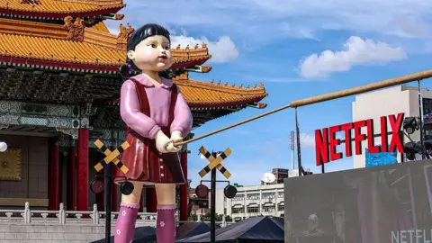 Getty Images A giant sculpture of a doll from the show Squid Game stands in front of a Korean temple, holding what appears to be a tug-o-war rope, next to a sign of the word Netflix