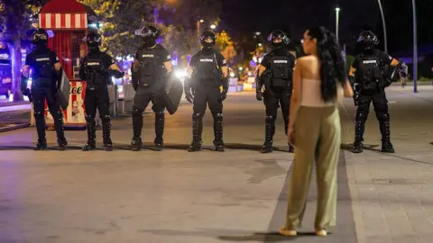 Getty Images A woman in a white top and green trousers stands in front of riot police dressed in black