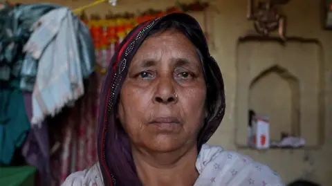 BBC A woman wearing a purple saree with her head covered looks into the camera 