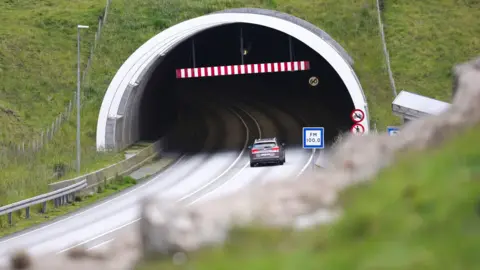 BBC A grey car enters a tunnel, driving past red "no pedestrian" and "no cyclist" roadsigns. The tunnel disappears into a grassy hillside. A blue and white radio station information sign reads "FM 100.0".