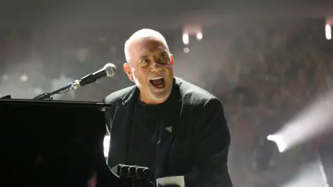Getty Images A bald man in a black t-shirt and black dinner jacket is performing on a piano, with a microphone pointed towards him. His mouth is open and smiling, with a crowd behind him, lit by spotlights.