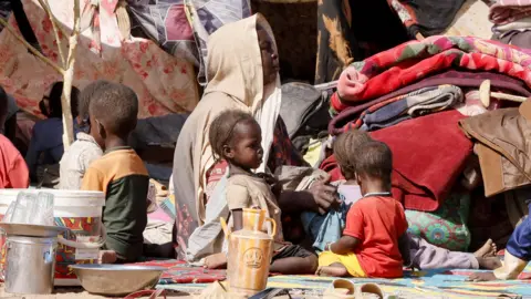Reuters A woman covered by a beige headscarf sits on a blanket, surrounded by five young children in colourful clothing, with braided hair. They are sitting amid piles of fabric, clothing and matting, under makeshift tents that have been propped up by small trees.