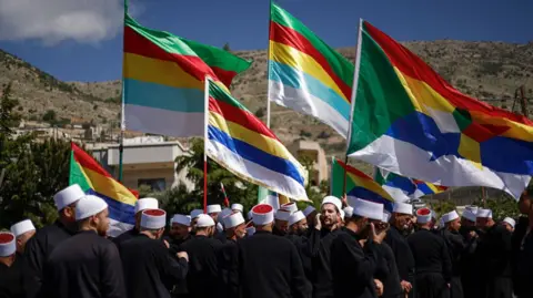 Getty Images Druze men lifting their flags in the air, wearing white hats and black robes