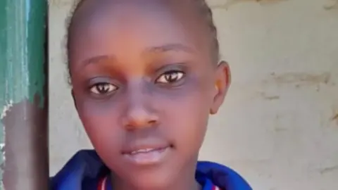 Njoki's family Bridgit Njoki stares at a camera, wearing a top with a blue collar. She stands in front of a white wall.