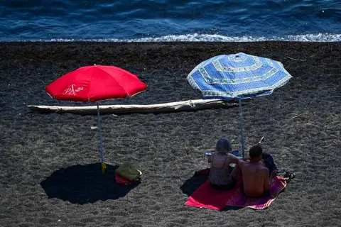 Getty Images A red and blue parasol on a beach next to the sea, which is at the top of the picture. Two beachgoers are resting in the shade under the blue parasol on the right.